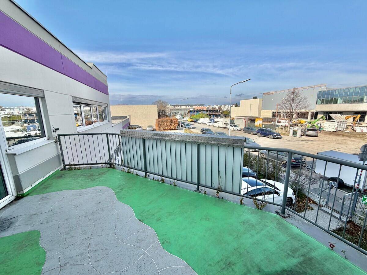 Balkon mit grün-grauem Boden und Metallgeländer, bietet Blick auf einen Parkplatz und Industriegebiet.