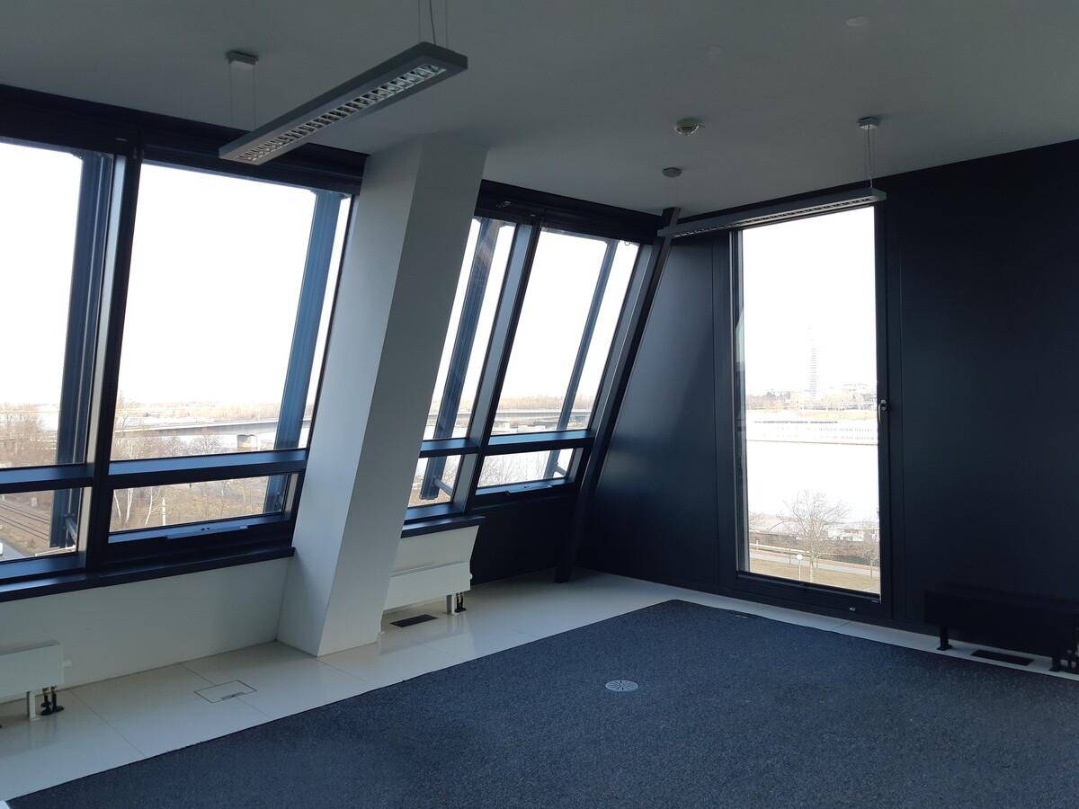 Leeres Büro mit schrägen Fenstern, dunklem Teppichboden und Blick auf das Wasser.