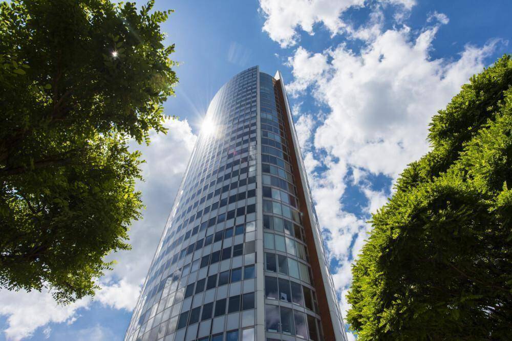 Beeindruckendes Hochhaus mit reflektierender Glasfassade, umgeben von Bäumen unter strahlend blauem Himmel.