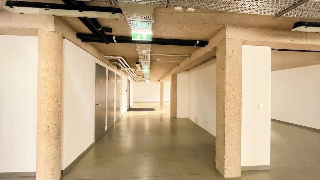 Langer Flur mit Betonstützen und Türen zu angrenzenden Räumen, beleuchtet durch Deckenleuchten.