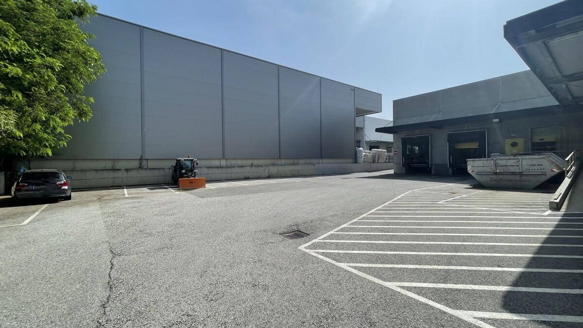 Großer Parkplatz neben einem modernen Industriegebäude mit Laderampen und Bäumen im Hintergrund.