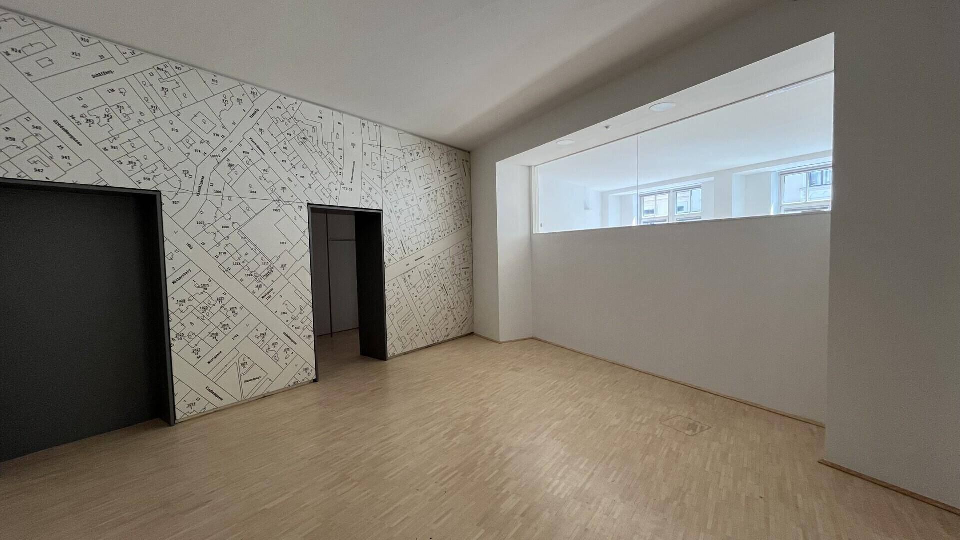 Geräumiger Büroraum mit hellem Holzfußboden, einer Wand mit Stadtplan-Muster und internem Fenster.