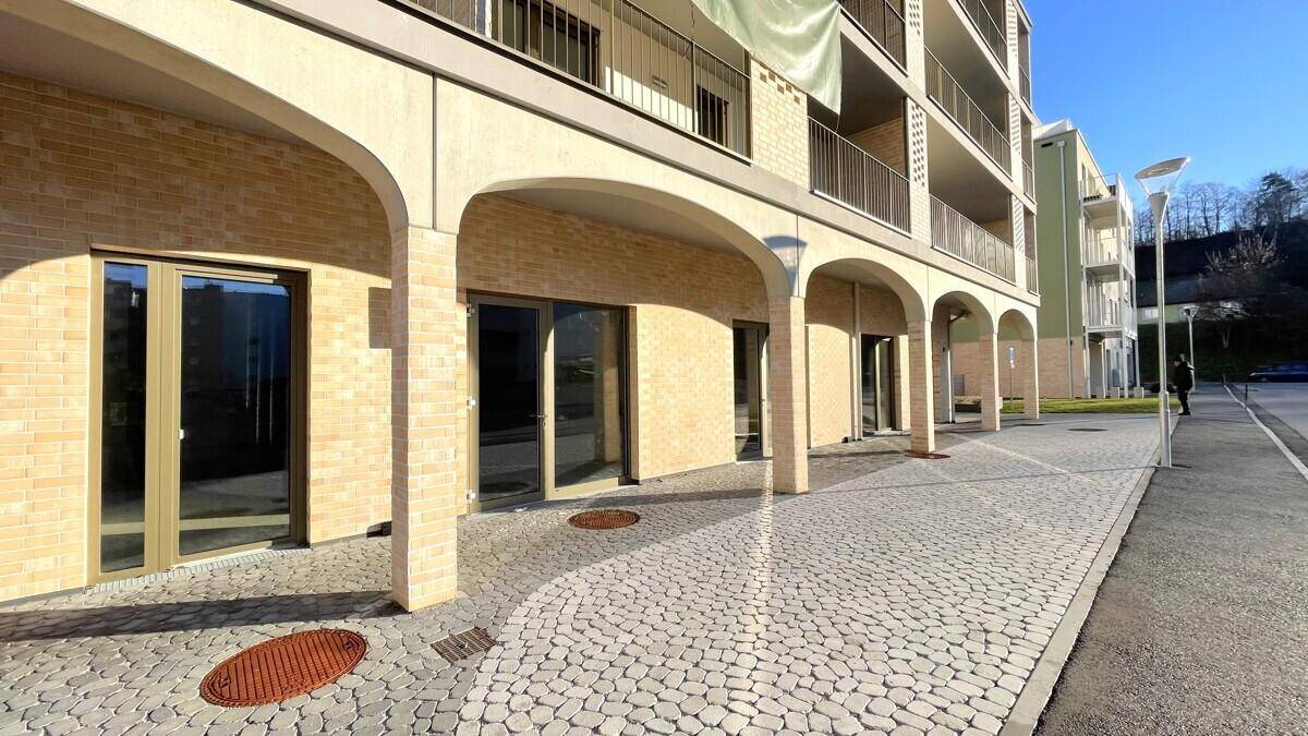 Seitenansicht eines modernen Gebäudes mit Arkaden, großen Fenstern und gepflastertem Gehweg unter blauem Himmel.