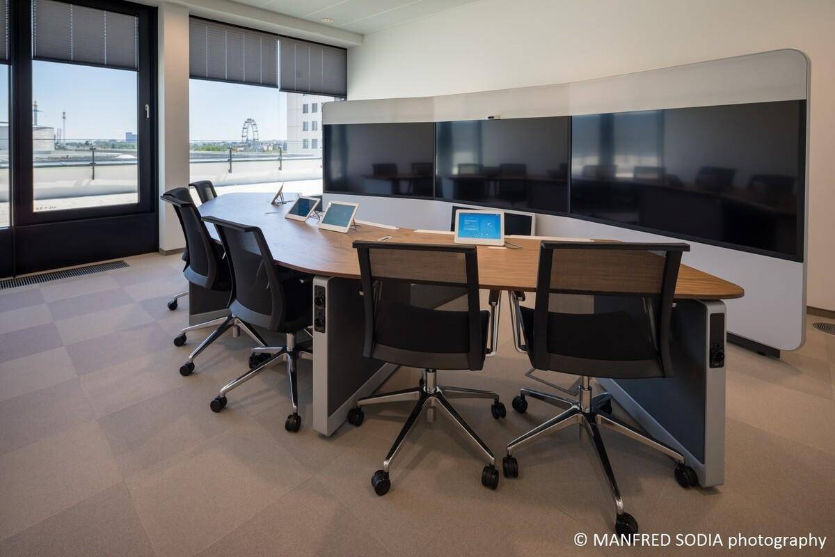 Moderner Konferenzraum mit ovalem Tisch, ergonomischen Stühlen und mehreren Bildschirmen.