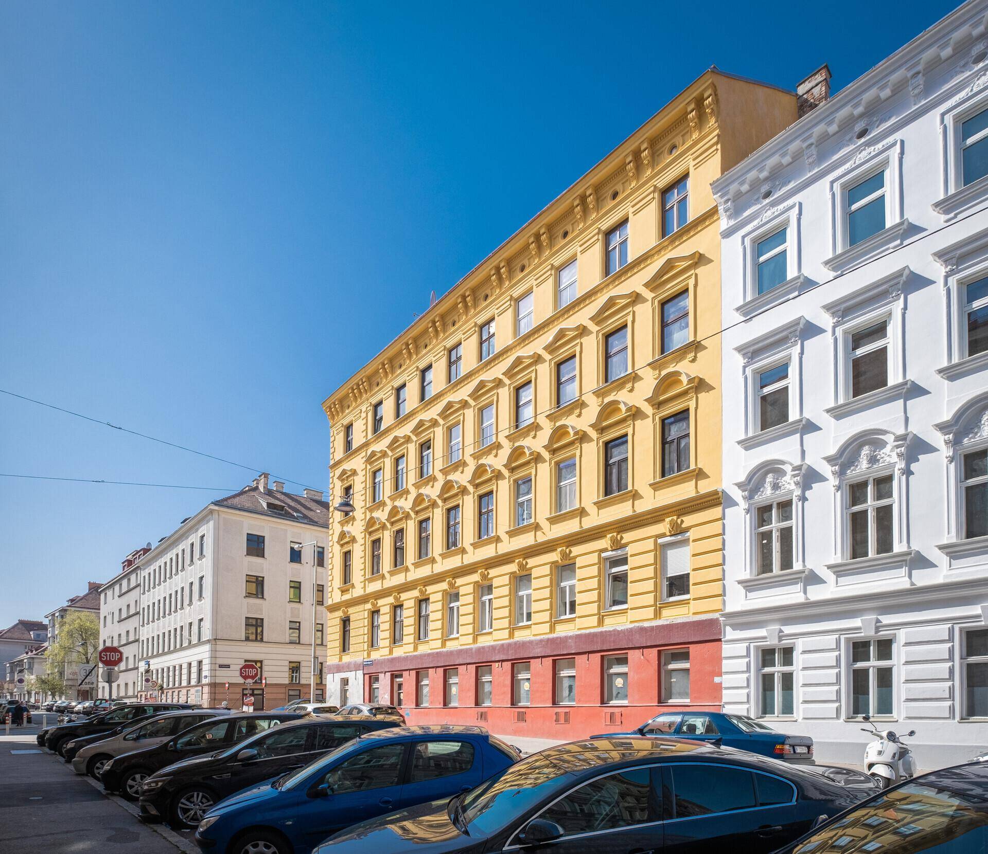 Ansicht der gelben Gebäudefassade entlang einer Straße mit parkenden Autos unter strahlend blauem Himmel.