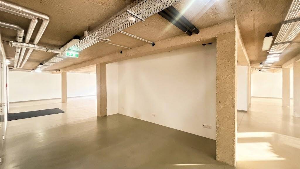 Offener Büroraum mit sichtbaren Deckeninstallationen und Betonstützen, der durch Fenster hell beleuchtet wird.