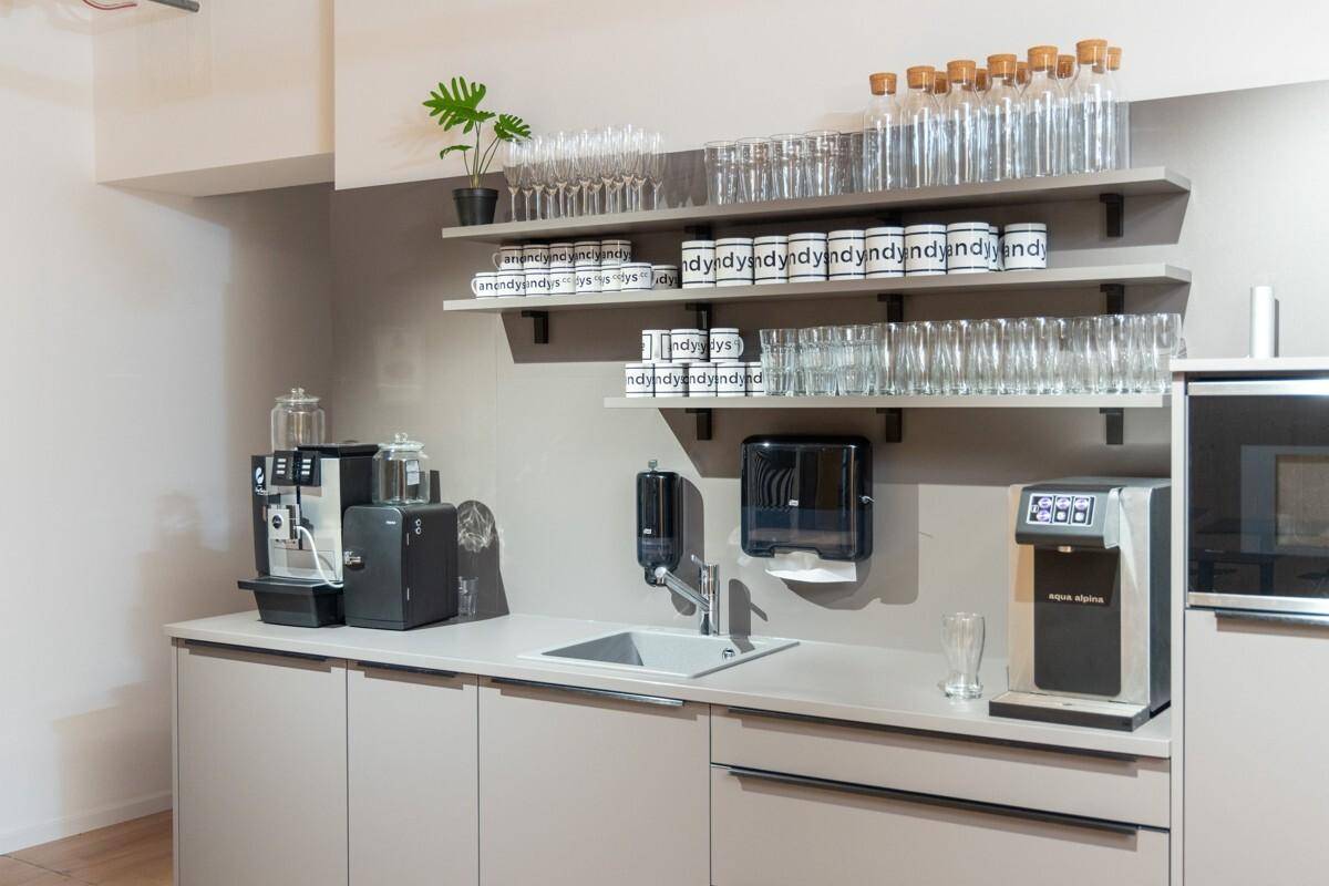 Funktionale Teeküche mit Spüle, Kaffeemaschine, Wasserspender und Regalen für Geschirr.