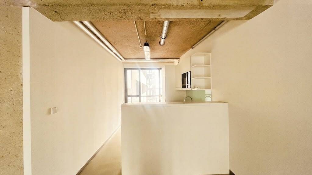 Kleiner Bürobereich mit einer Theke, Regalen und einem Fenster, das Tageslicht hereinlässt. Die Decke ist unverputzt.