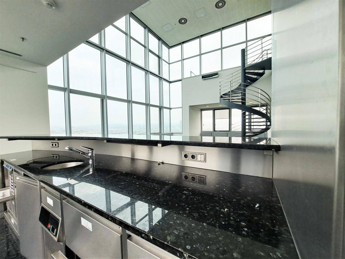 Moderne Küchenzeile mit dunkler Granitarbeitsplatte und Edelstahlfronten, im Hintergrund die großen Fenster und Wendeltreppe.