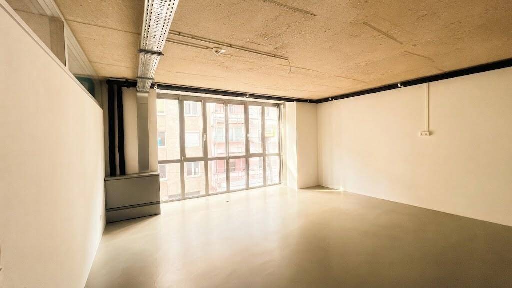 Eckbüro mit großen Fenstern, die viel Tageslicht bieten, und einer unverputzten Decke mit sichtbaren Installationen.