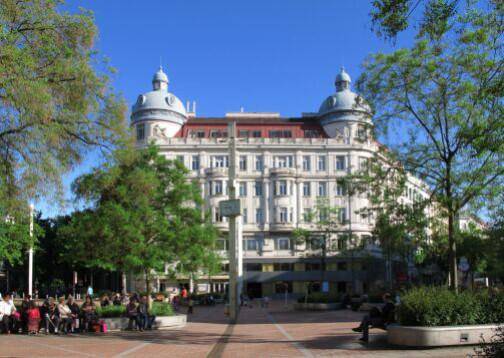 Historisches Gebäude mit markanten Kuppeln und einer belebten Platzanlage mit Bäumen im Vordergrund.