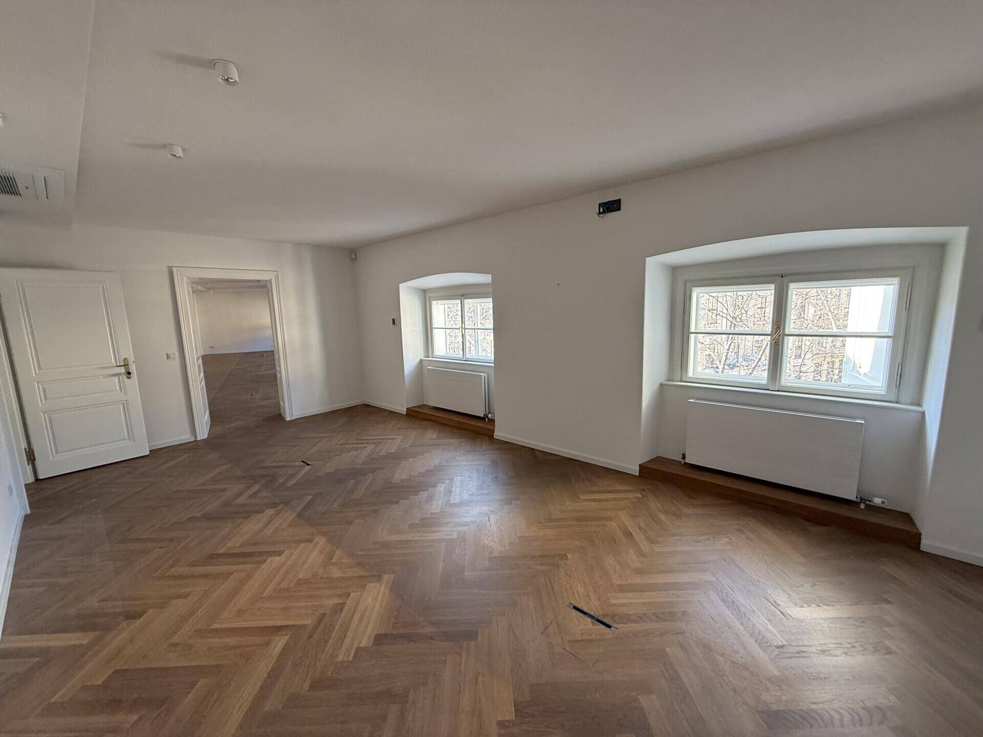 Geräumiger Raum mit Fischgrätparkett, weißen Wänden und klassischen Fenstern, die den Blick nach draußen freigeben.