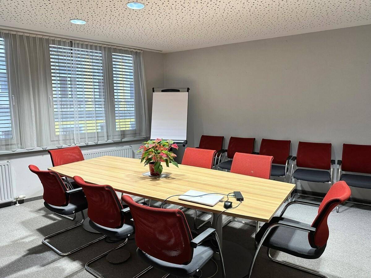 Konferenzraum mit großem Holztisch, roten Stühlen und Teppichboden, ideal für Besprechungen.