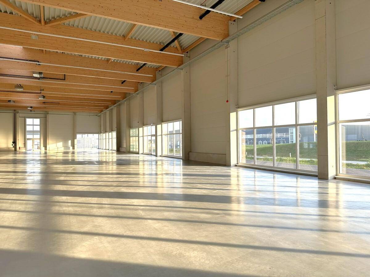 Geräumige, helle Lagerhalle mit hohen Decken, Holzbalken und großen Fenstern, die viel Tageslicht hereinlassen.