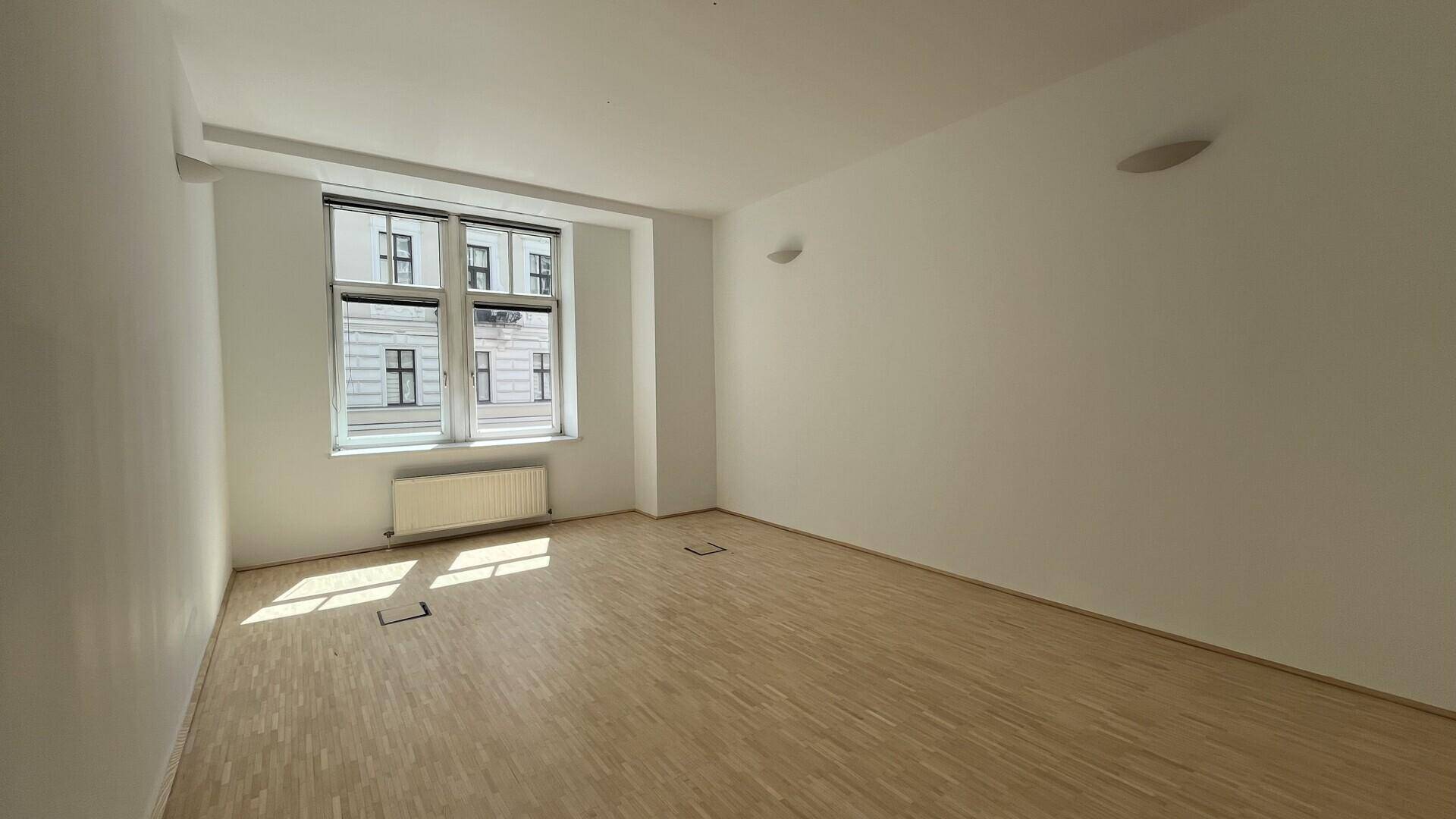 Leerer Büroraum mit hellem Holzfußboden, einem großen Fenster und Heizkörper, bereit zur individuellen Gestaltung.