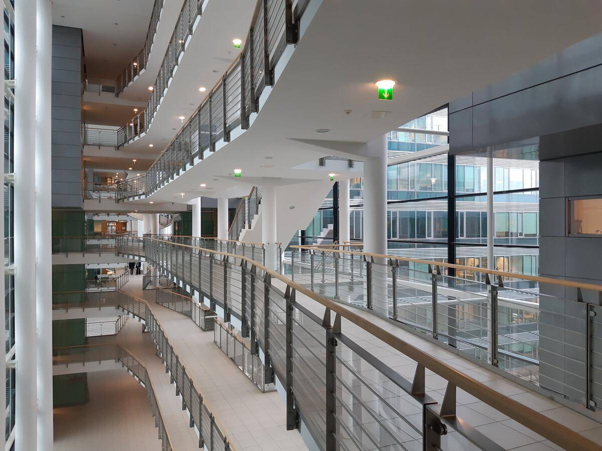 Heller, mehrstöckiger Atrium-Flur in einem modernen Bürogebäude mit Glasgeländern und viel Tageslicht.