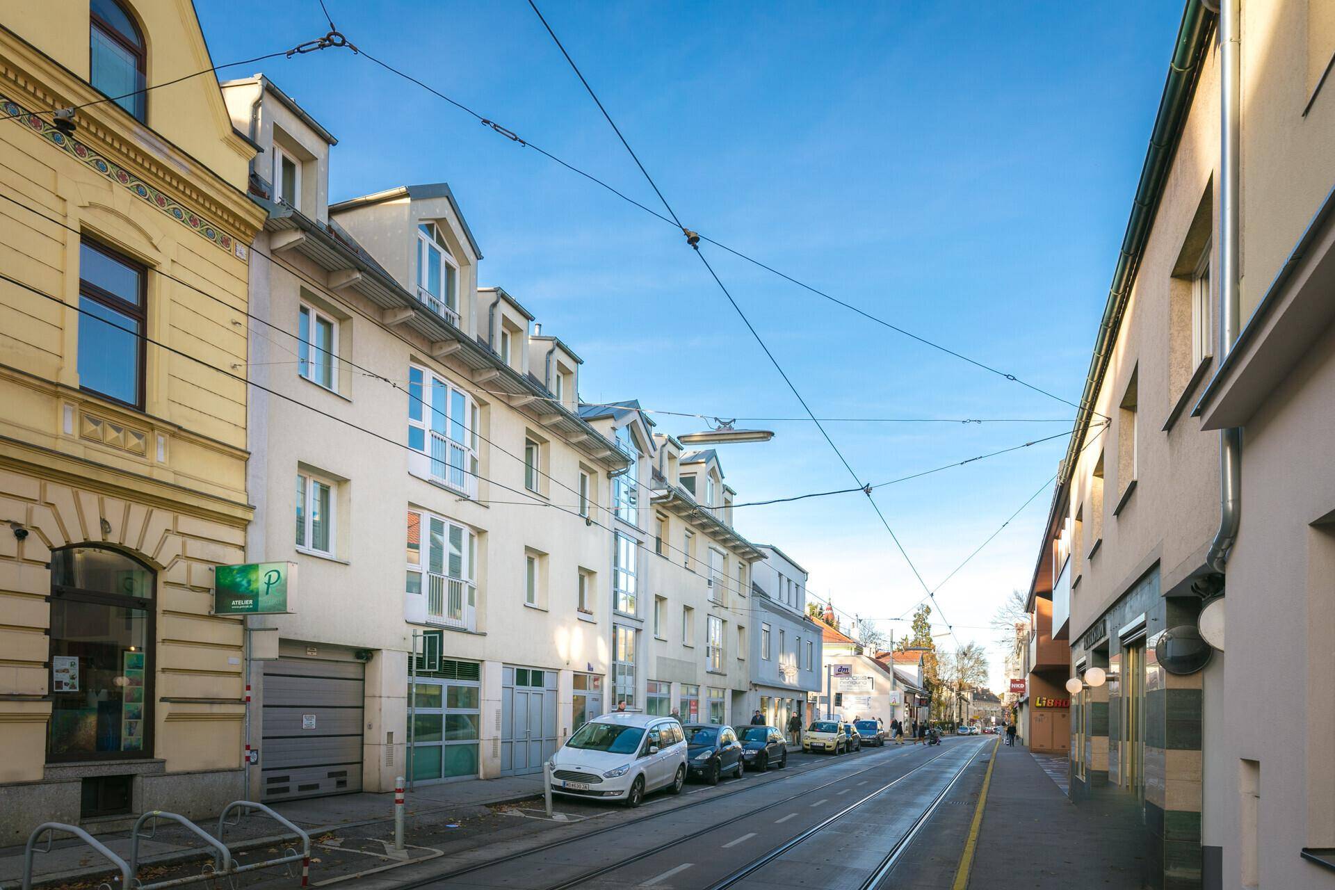 Seitenansicht des Gebäudes an einer belebten Straße mit Straßenbahnschienen und parkenden Autos.