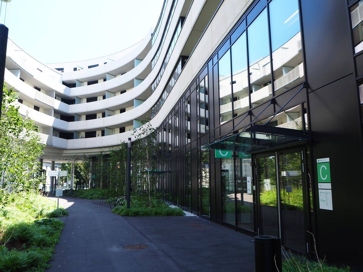 Geschwungene moderne Gebäudefassade mit großen Glasflächen und einem begrünten Außenbereich.