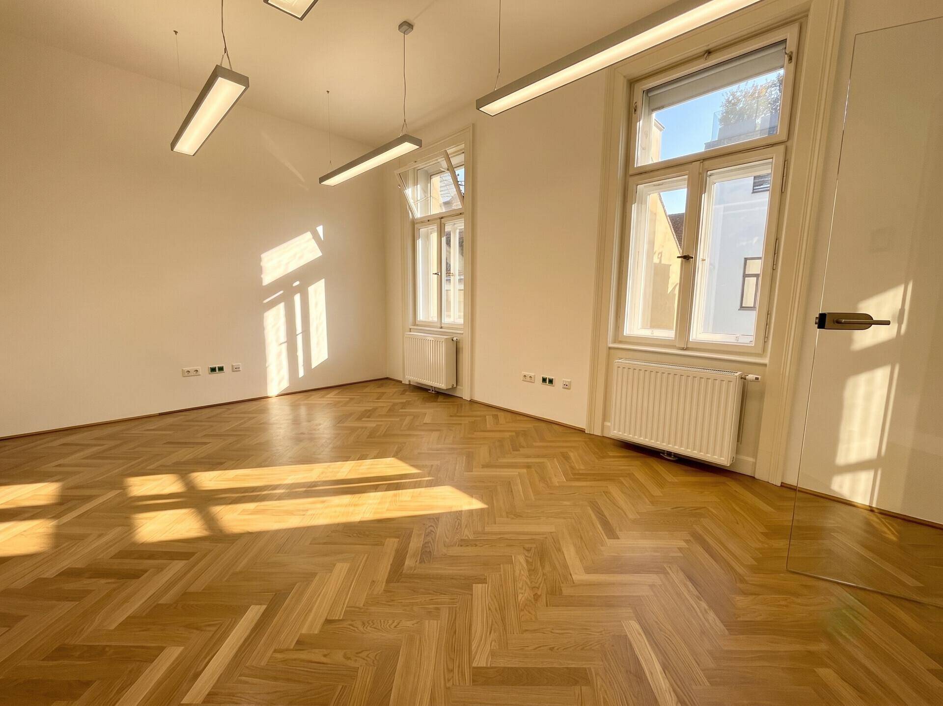 Helles Büro mit Fischgrätparkett, großen Fenstern und einer modernen Glastür, die den Raum öffnet.