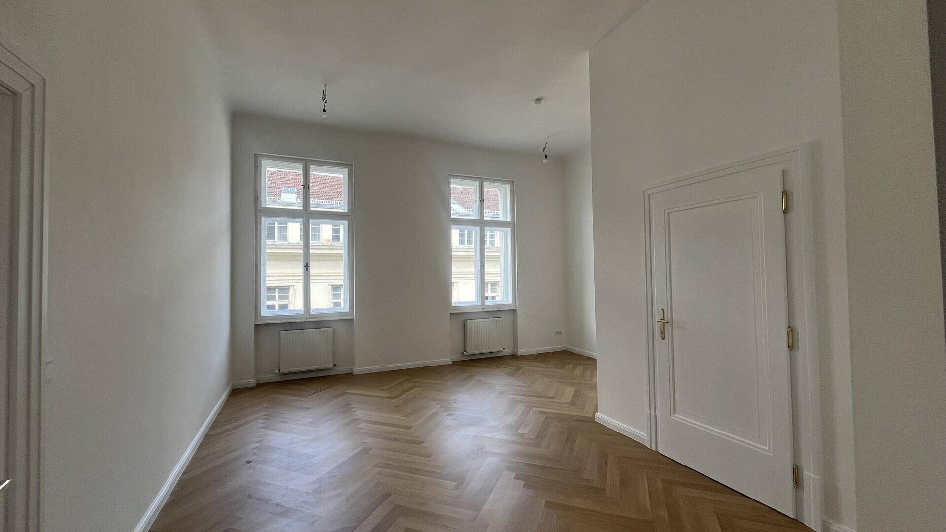 Geräumiger, heller Raum mit Fischgrätparkett, weißen Wänden und klassischen Türen, bereit zur Einrichtung.