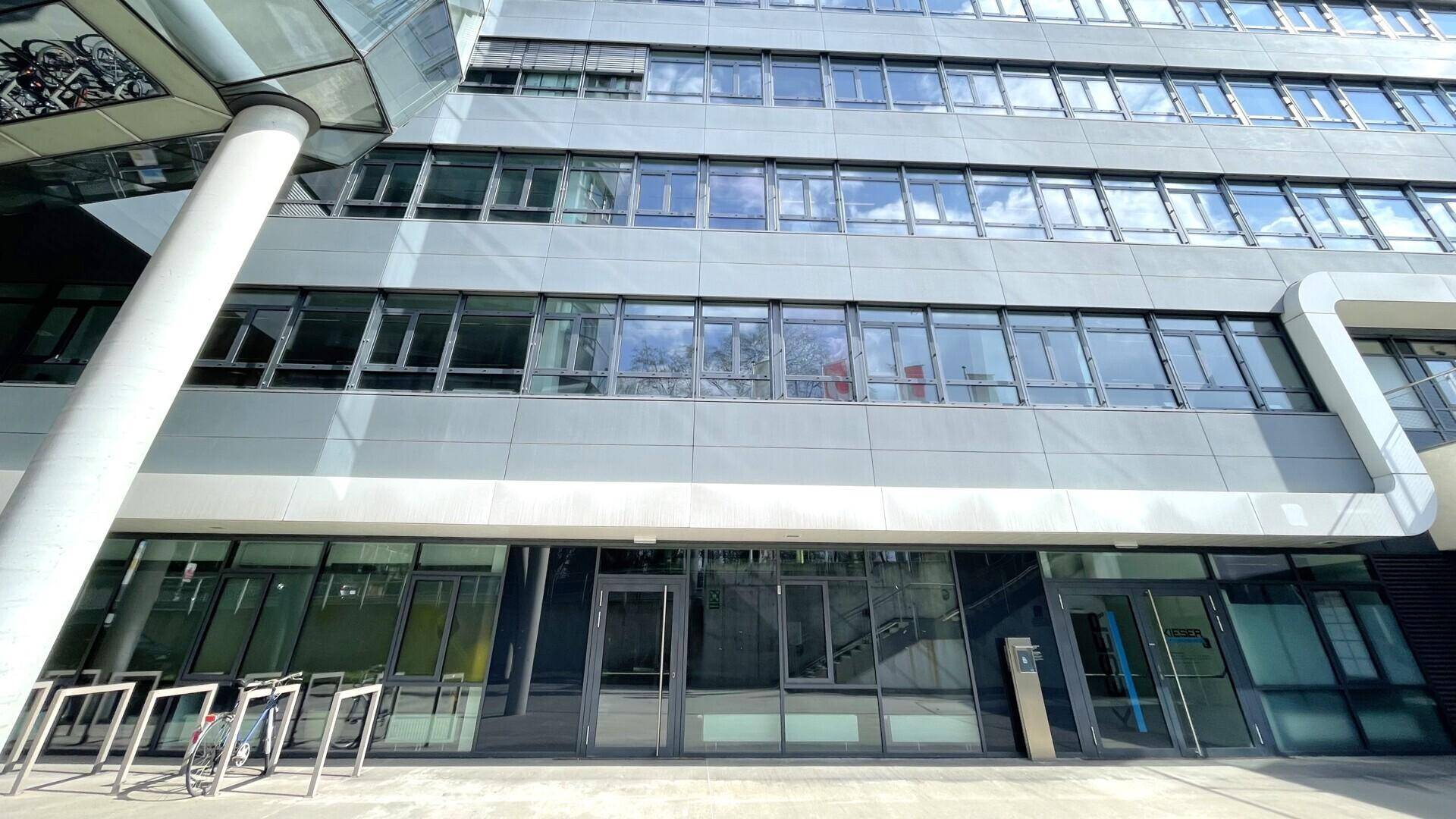 Nahaufnahme der modernen Bürogebäude-Fassade mit großen Fenstern und dem Haupteingangsbereich im Sonnenlicht.