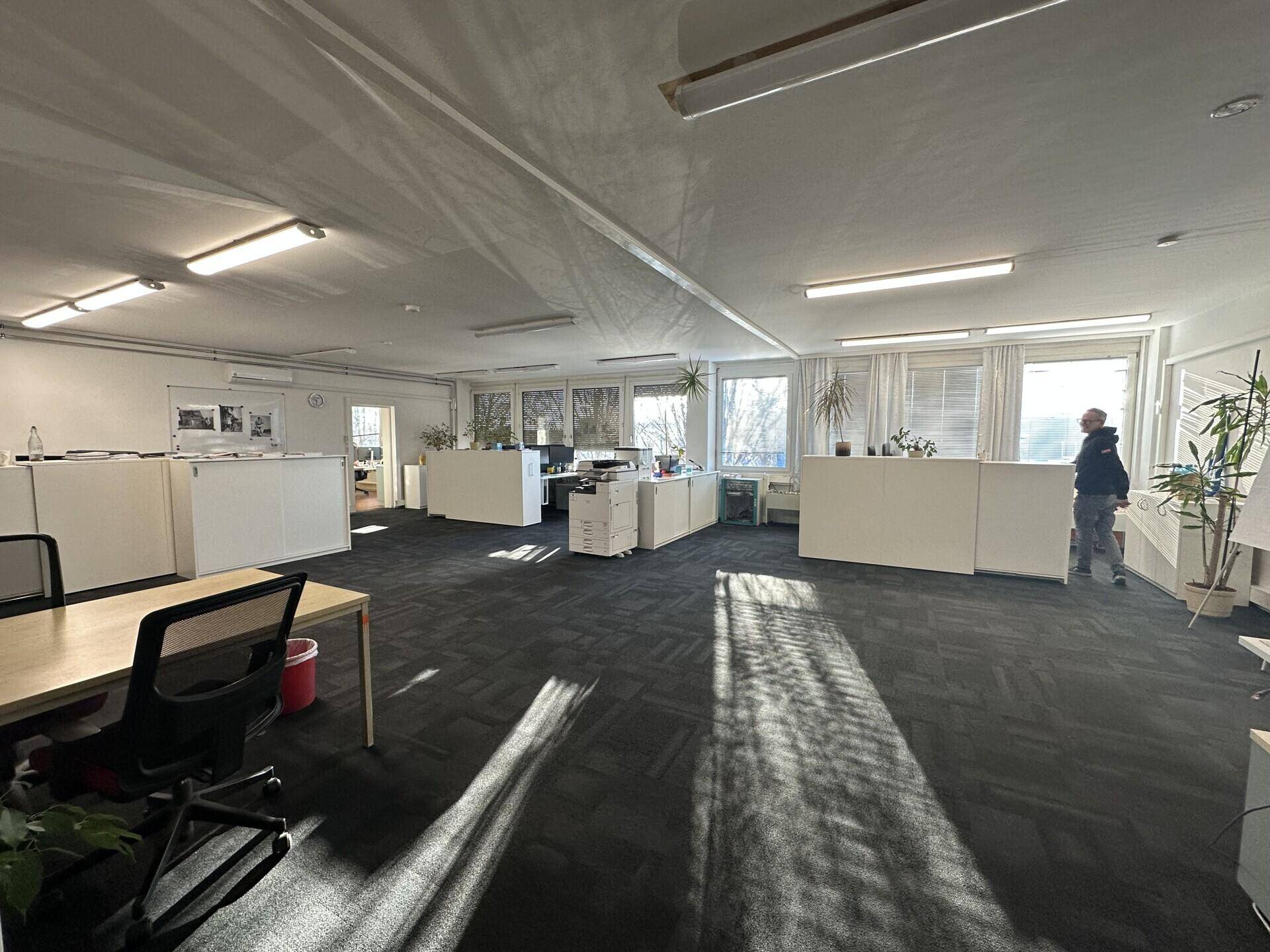 Großes Großraumbüro mit mehreren Arbeitsplätzen, Teppichboden und viel Tageslicht.