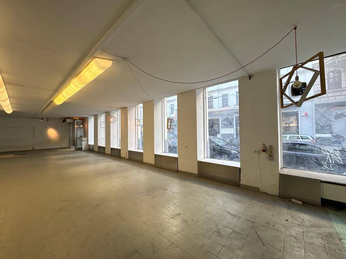 Ein leerstehender, langer Gewerberaum mit großen Fenstern zur Straße, der viel Tageslicht hereinlässt.