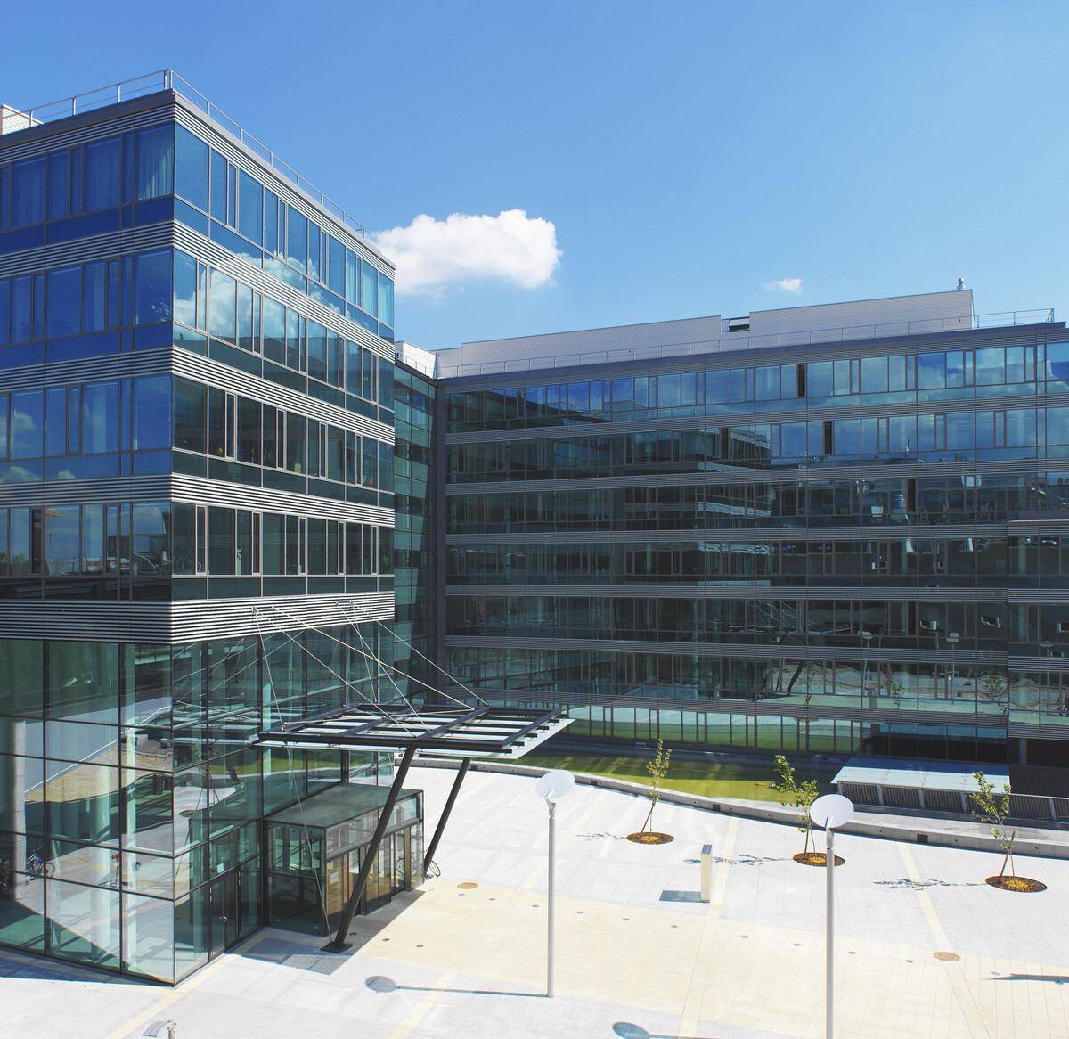 Ansicht eines modernen Bürokomplexes mit weitläufiger Glasfassade, einem eleganten Eingangsbereich und Außenparkplätzen.
