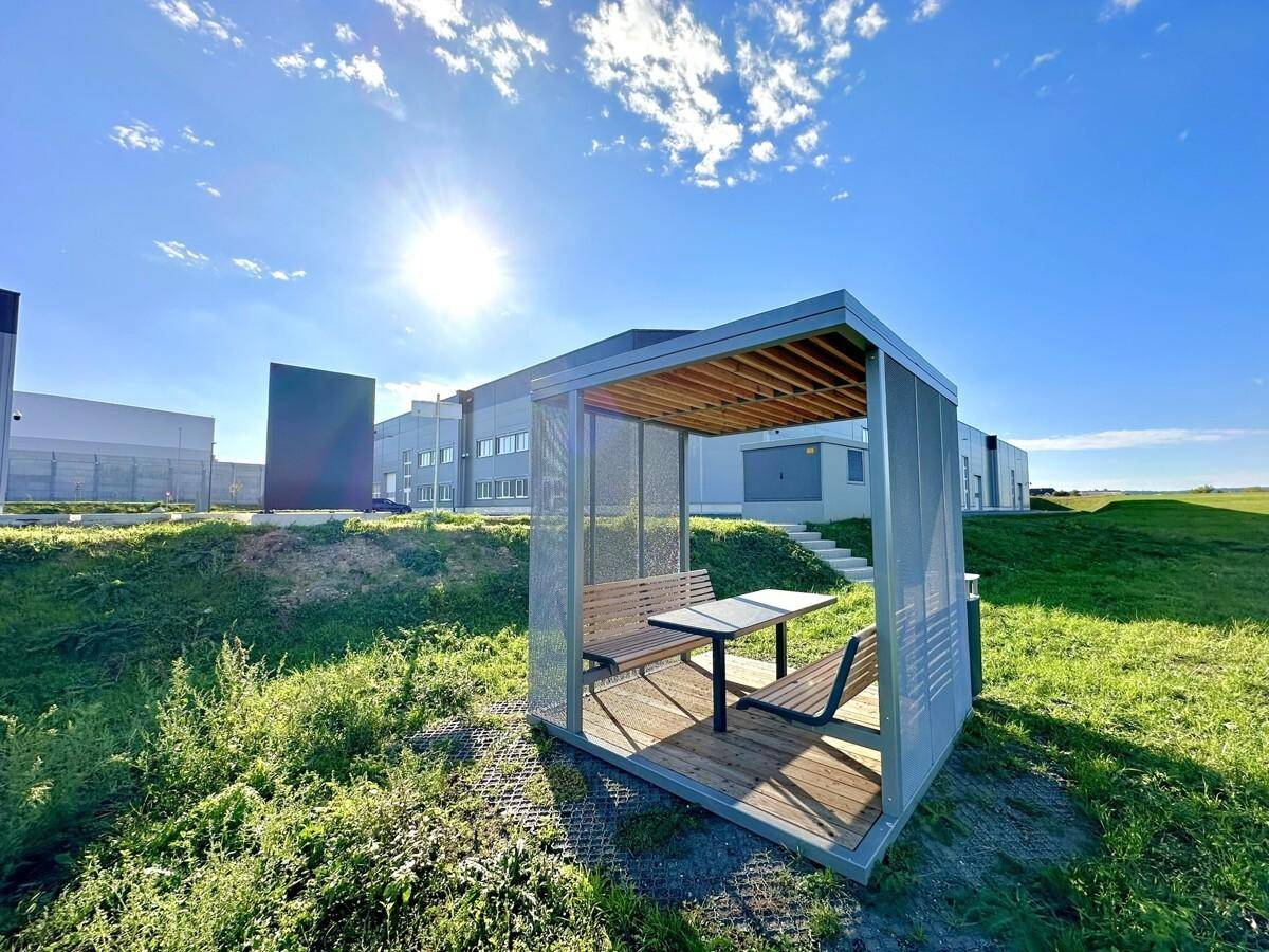 Moderner Pavillon mit Sitzgelegenheiten in einer grünen Umgebung unter blauem Himmel und Sonnenschein.