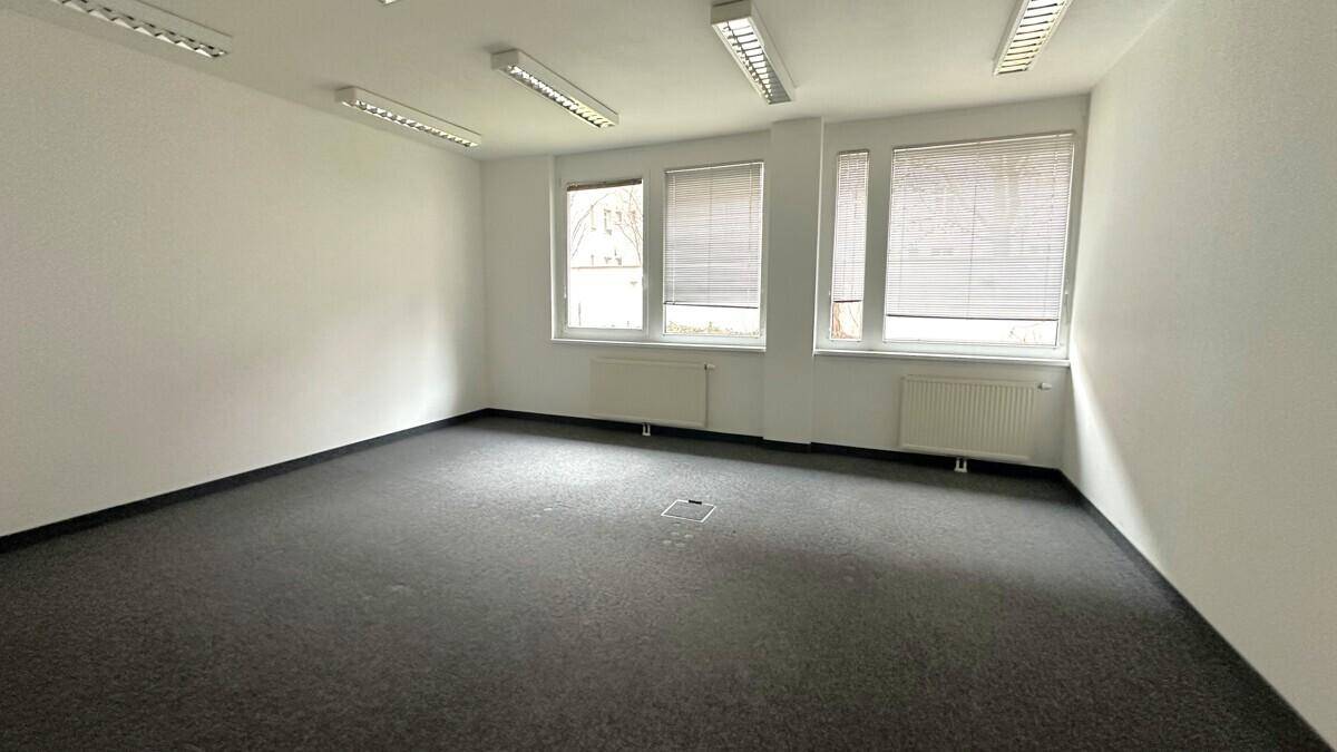 Leerer Büroraum mit grauem Teppichboden, weißen Wänden und Jalousien an den Fenstern für Privatsphäre.