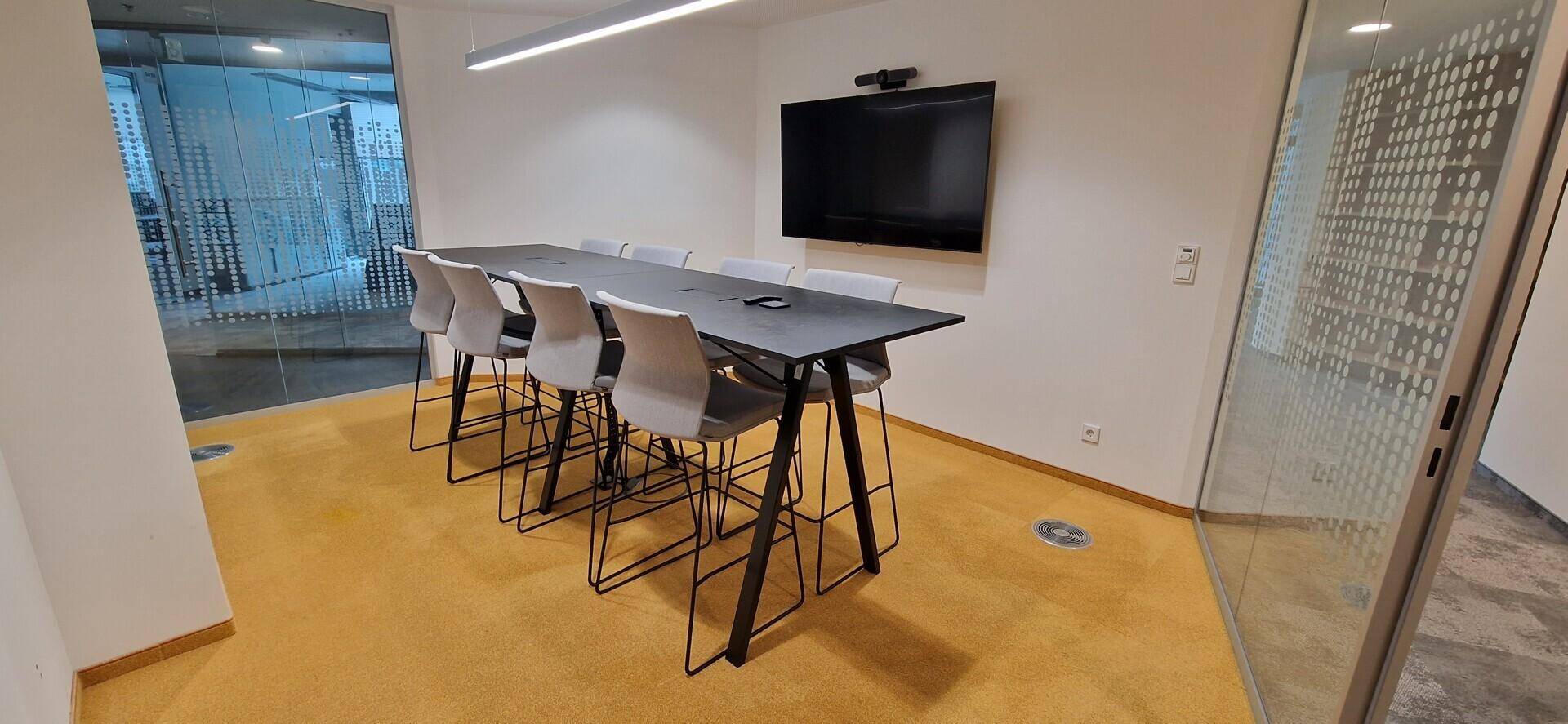Moderner Konferenzraum mit langem Tisch, Barhockern, Wandfernseher und gelbem Teppichboden.