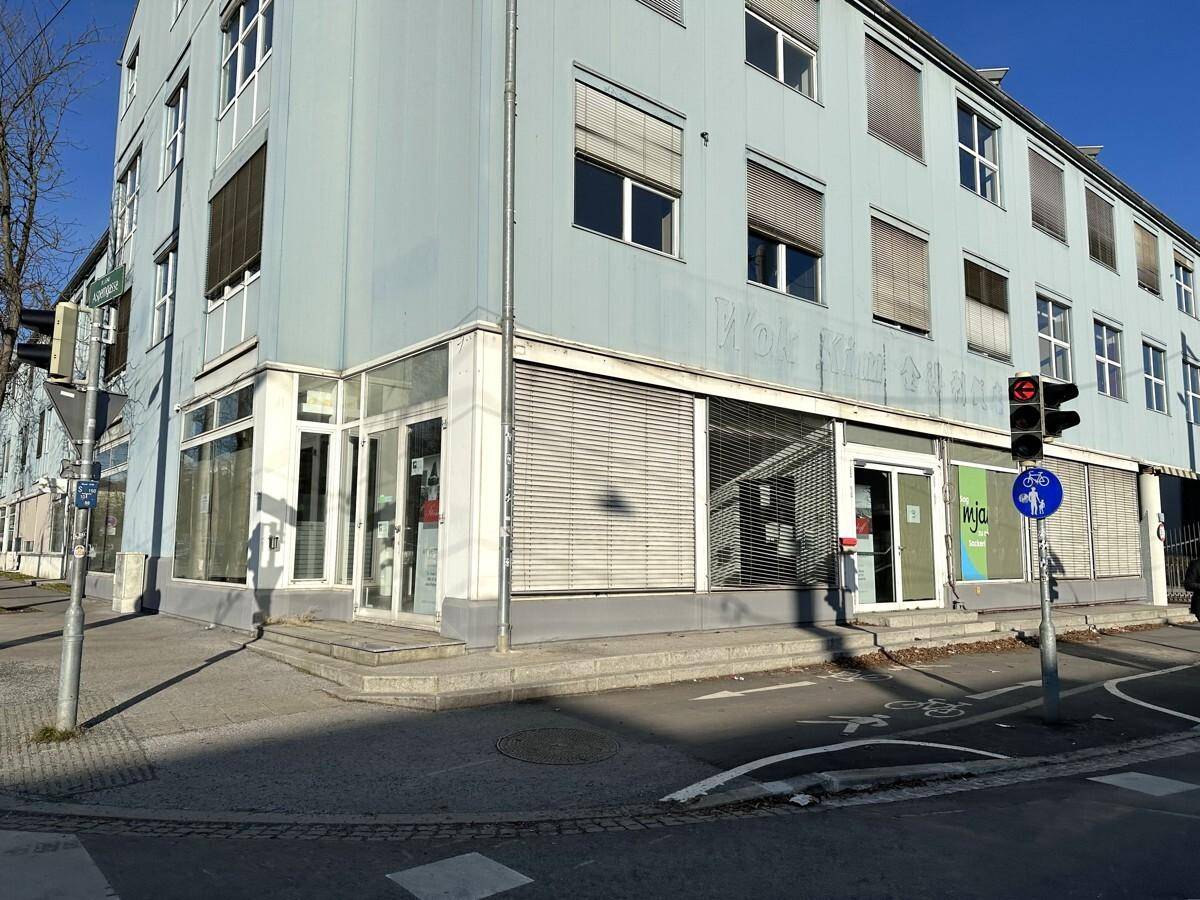 Gebäudeansicht an einer Straßenecke mit Geschäften im Erdgeschoss und Rollläden an den Fenstern.