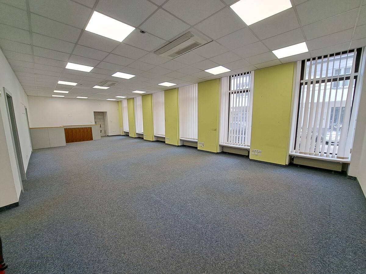 Großer, leerer Büroraum mit Teppichboden, vielen Fenstern und abgehängter Decke mit integrierter Beleuchtung.