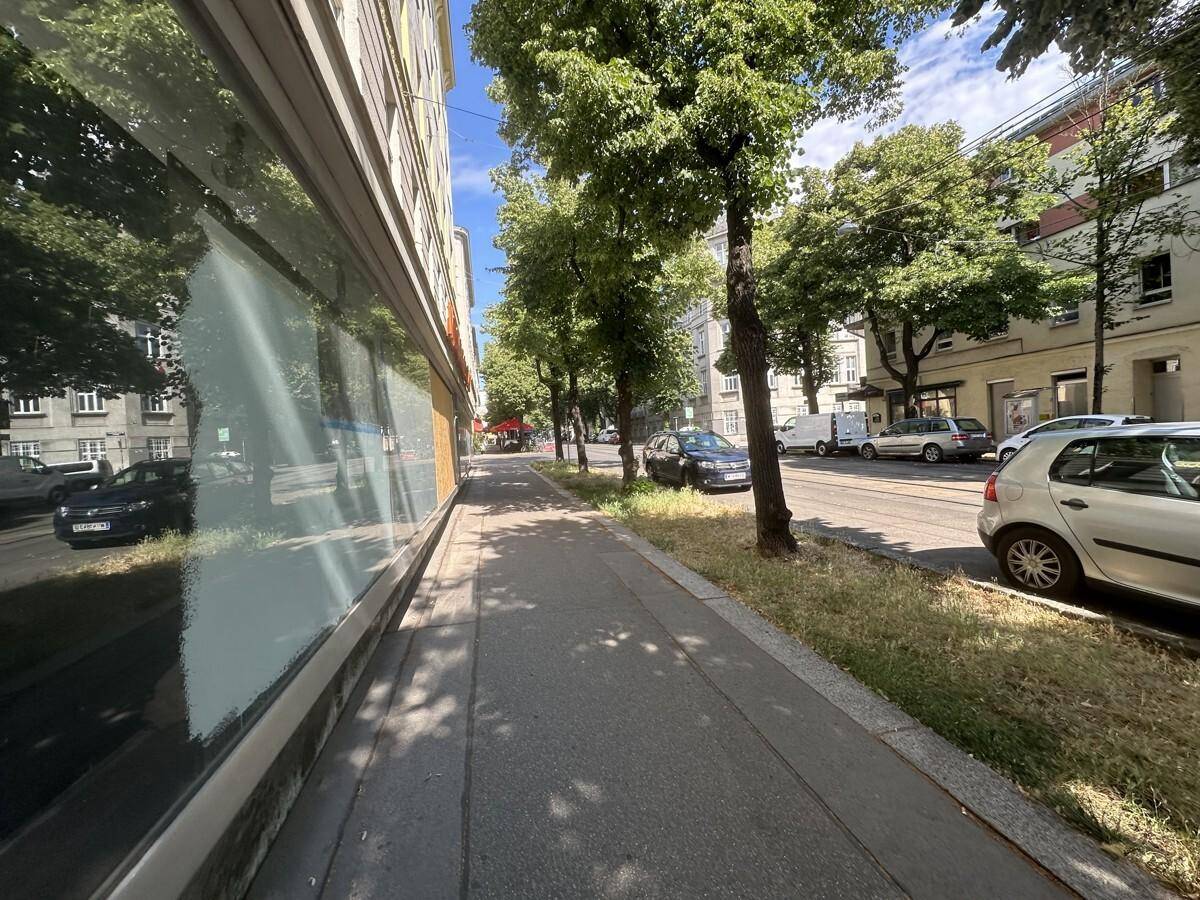 Eine lange Perspektive des Bürgersteigs entlang der Gebäudefassade mit Bäumen und parkenden Autos.