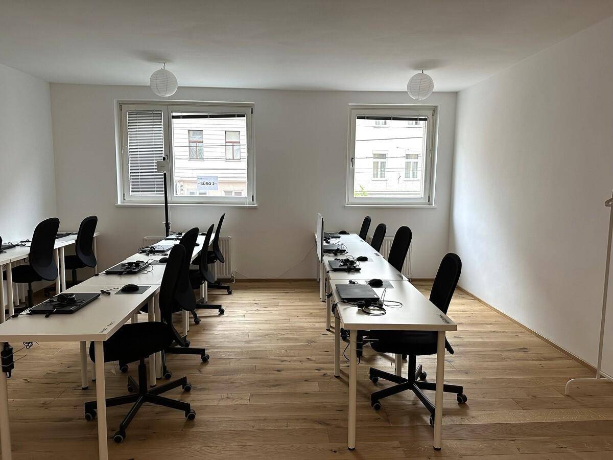 Funktionaler Büroraum mit mehreren Arbeitsplätzen, Holzboden und Fenstern mit Jalousien.