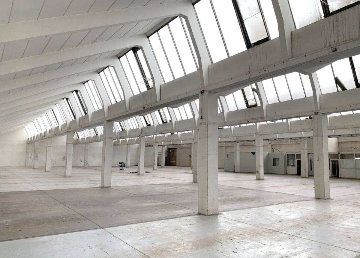 Geräumige Lagerhalle mit hohen Decken, vielen Stützpfeilern und großen Dachfenstern, die viel Tageslicht hereinlassen.