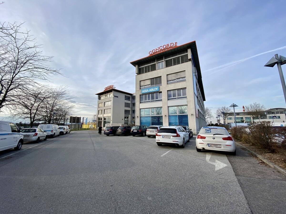 Ansicht eines modernen Bürokomplexes mit Parkplätzen und Bäumen unter einem leicht bewölkten Himmel.
