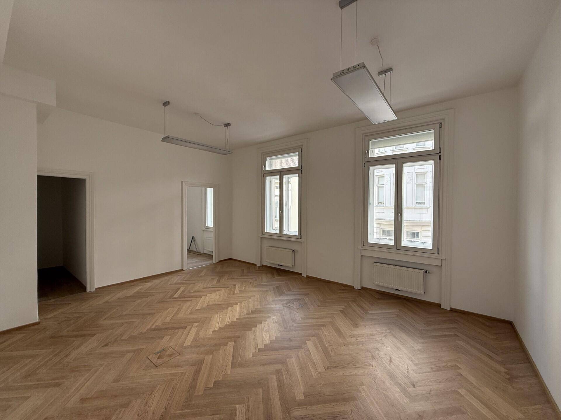 Leeres Büro mit Fischgrätparkett, zwei großen Fenstern und Zugang zu weiteren Räumen, hell und geräumig.