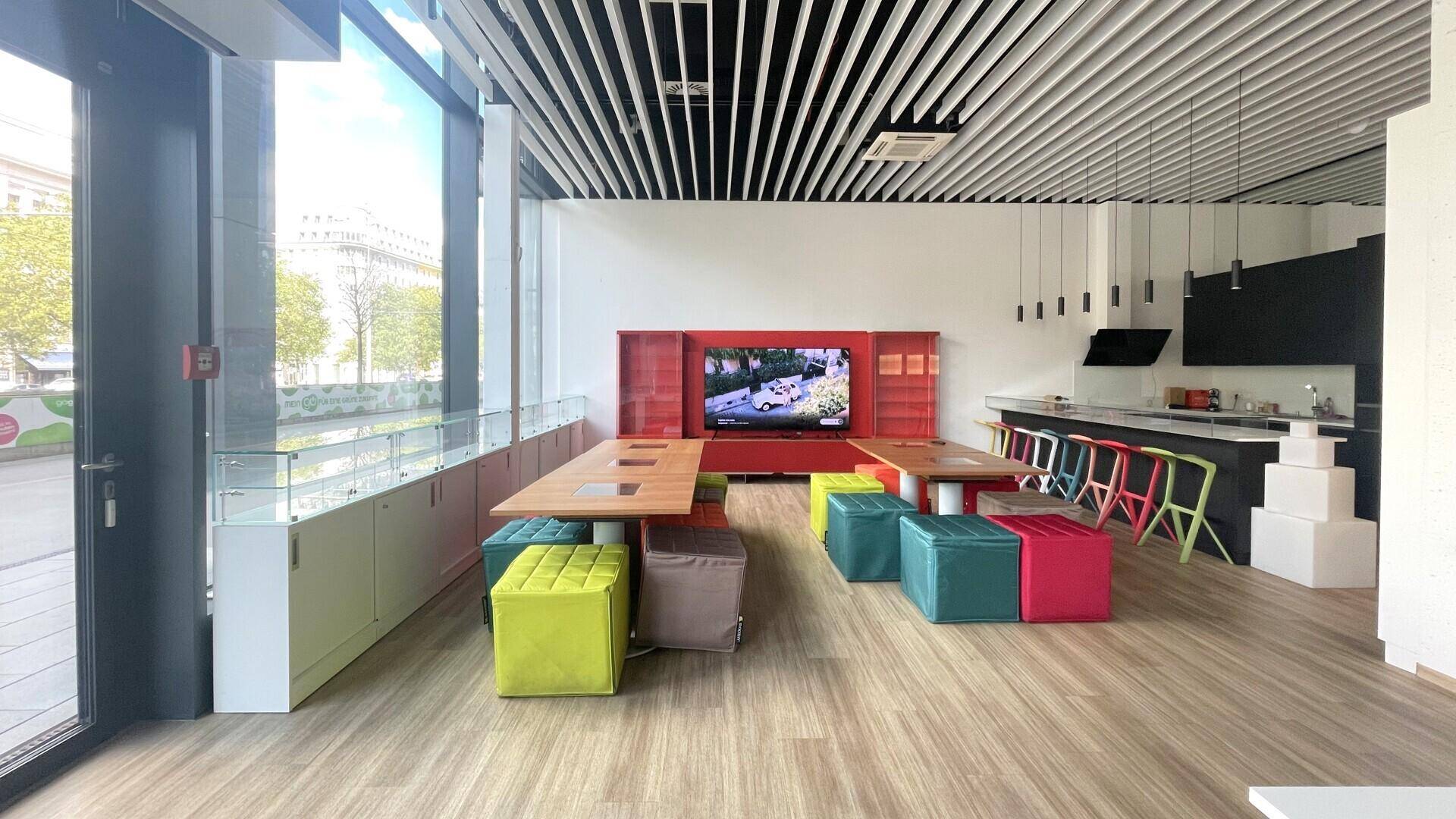Geräumiger Verkaufsraum mit Holzboden, bunten Sitzwürfeln und einer integrierten Küchenzeile im Hintergrund.