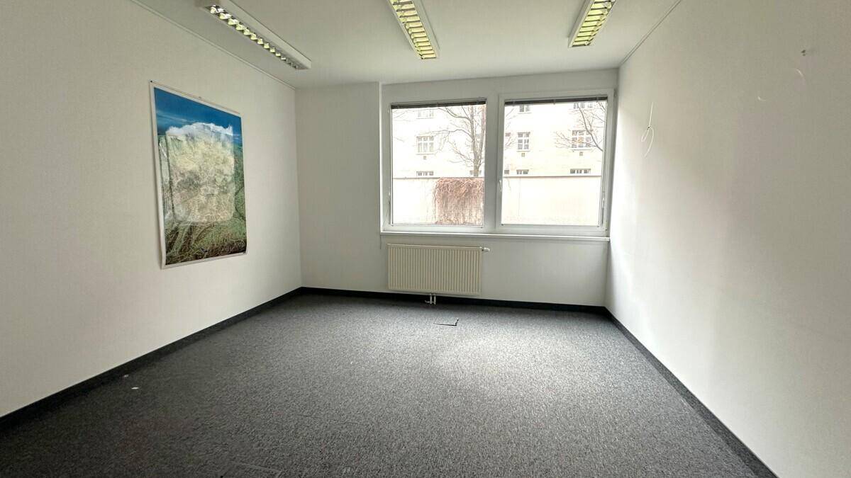 Leeres Büro mit grauem Teppichboden, weißen Wänden und einem Bild an der Wand, gut beleuchtet.