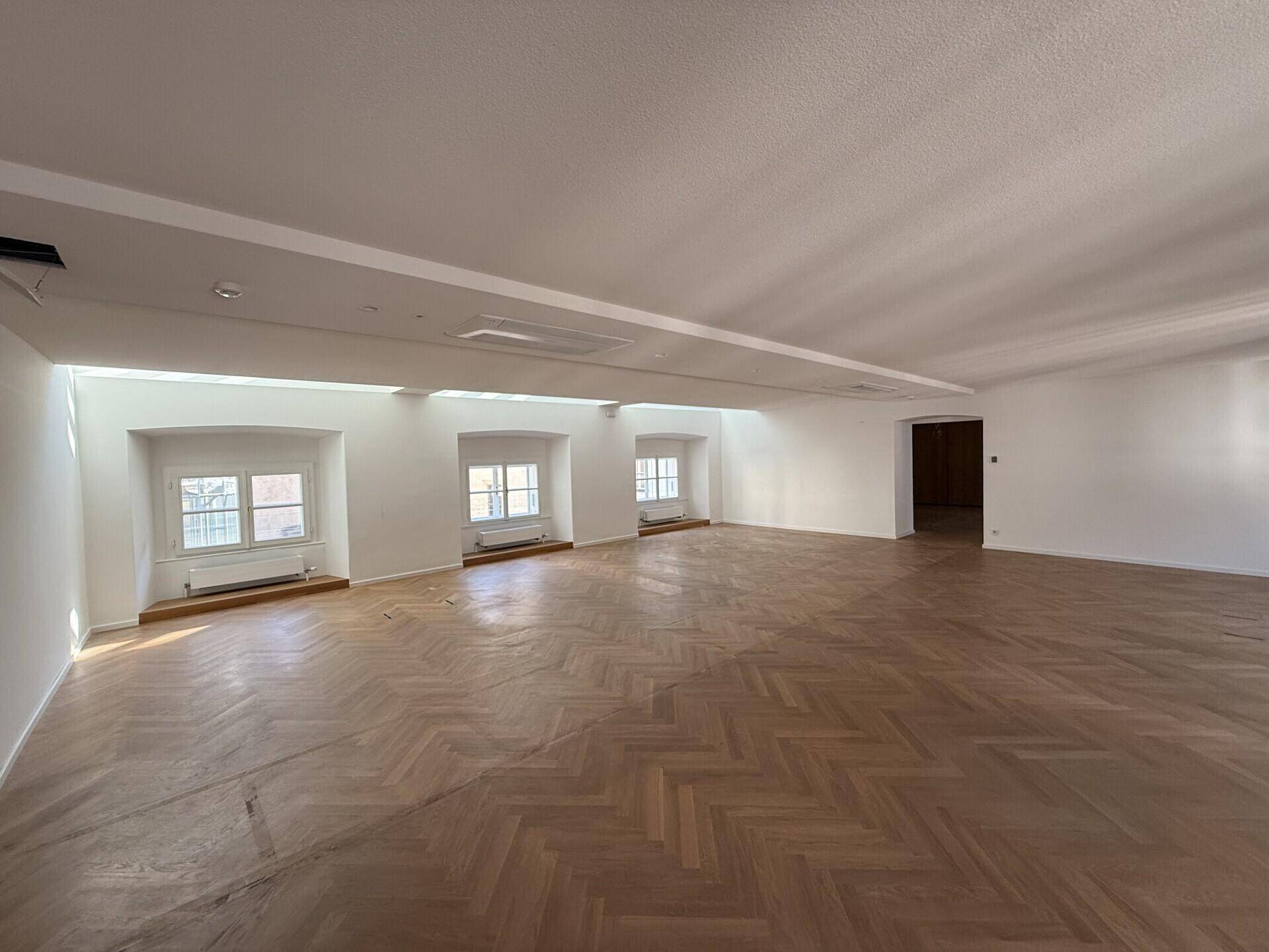 Geräumiger, heller Raum mit Fischgrätparkett, weißen Wänden und tiefen Fensternischen, die viel Tageslicht hereinlassen.