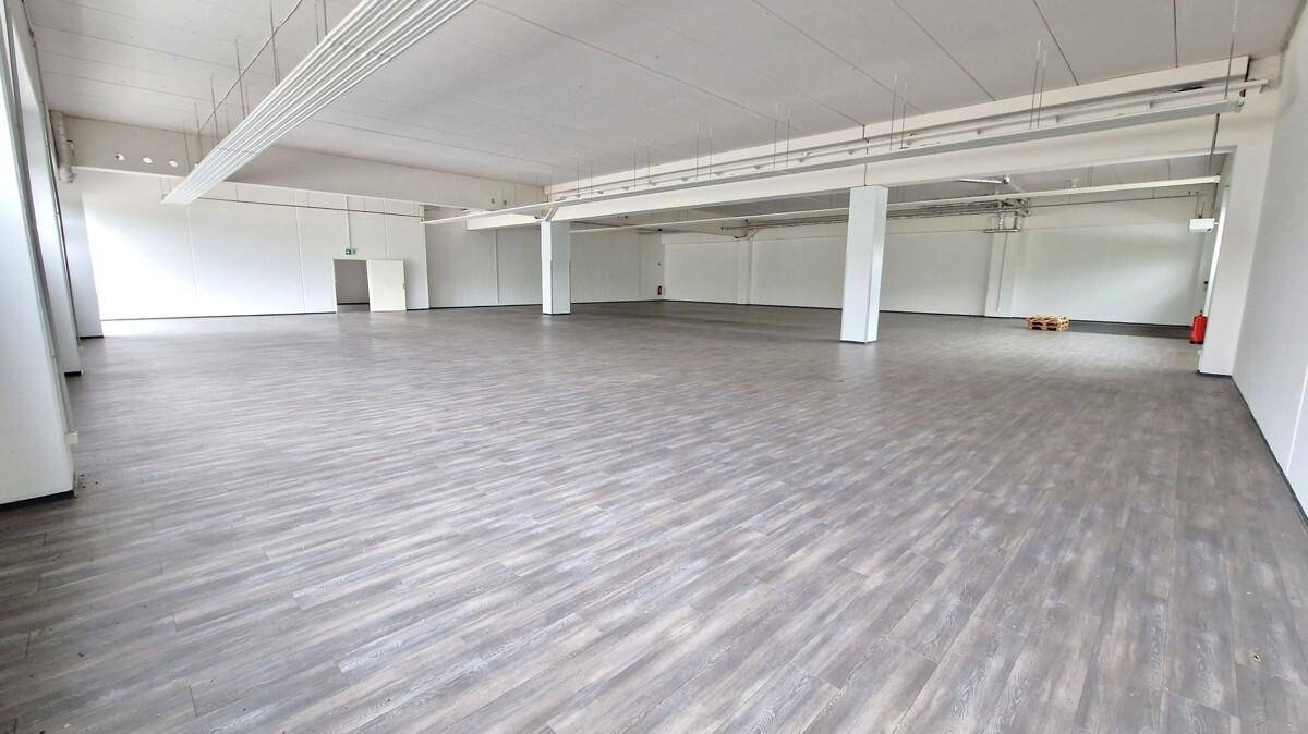 Geräumiger, leerer Gewerberaum mit grauem Boden, weißen Wänden und sichtbaren Deckenbalken und Beleuchtung.
