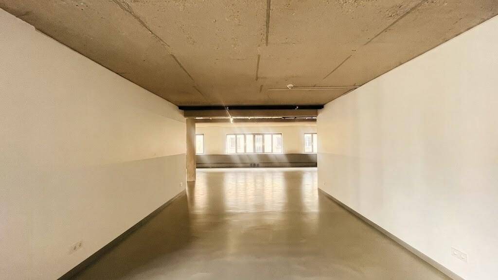 Langer, heller Flur mit weißen Wänden und Betonboden, der zu einem offenen Bereich mit Fenstern führt.