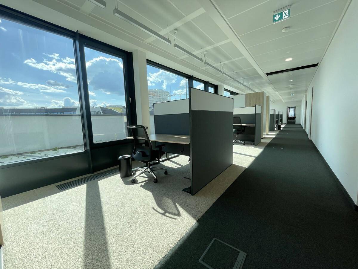 Moderner Bürogang mit Arbeitsplätzen entlang der Fensterfront und Teppichboden.