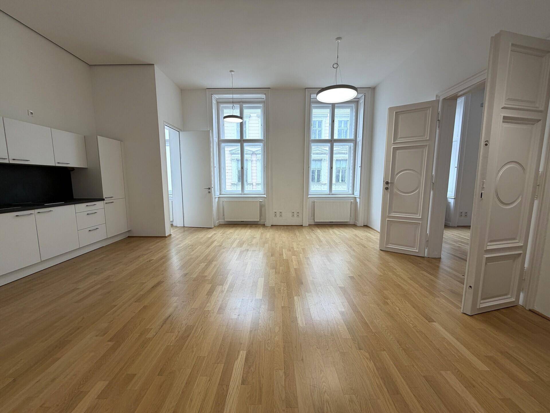Heller Raum mit Holzparkett, hohen Decken und einer modernen Küchenzeile im Hintergrund.
