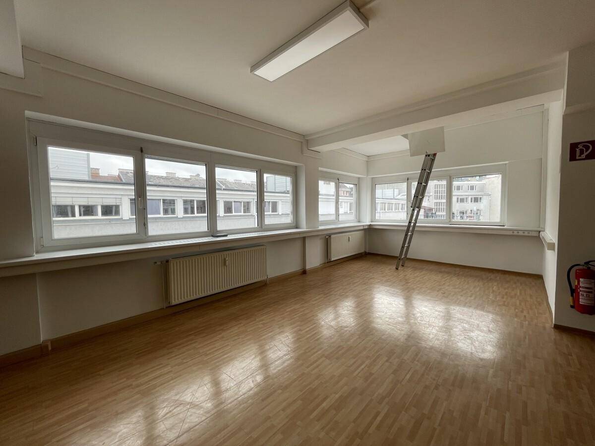 Leerer Büroraum mit hellem Holzfußboden, großen Fenstern und einer Leiter, die an der Wand lehnt.