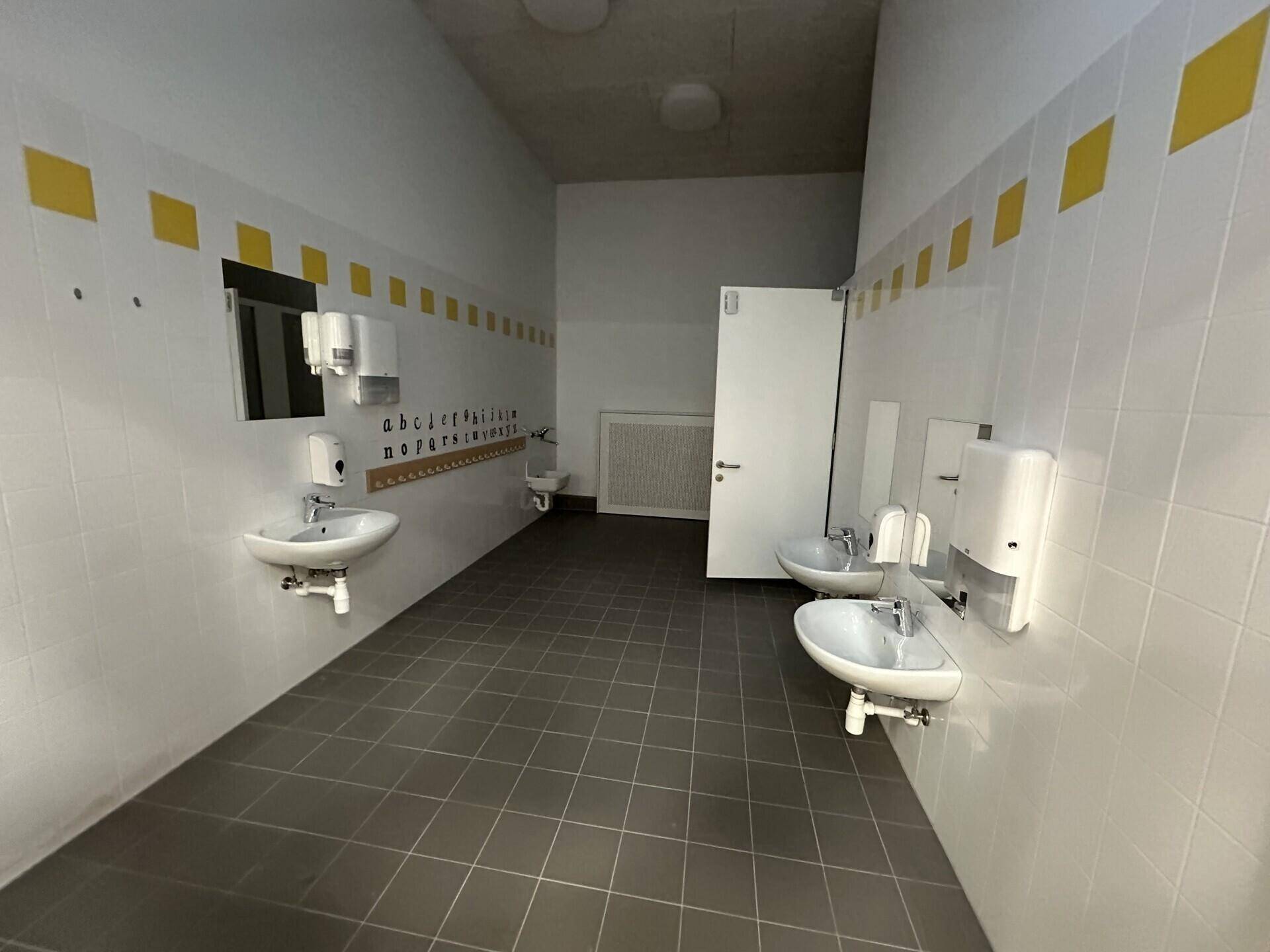 Geräumiger Waschraum mit mehreren Waschbecken, Fliesenboden und gelben Akzenten an den Wänden.