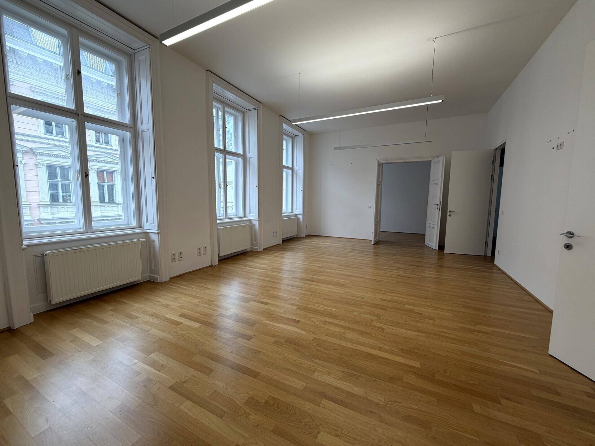 Geräumiges Büro mit Holzboden, hohen Fenstern und weiß gestrichenen Wänden.