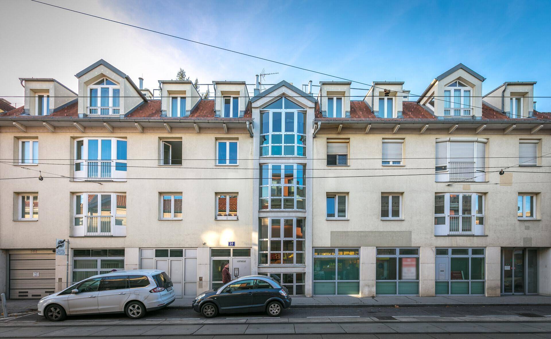 Ansicht der beigen Gebäudefassade mit vielen Fenstern, Dachgauben und einem verglasten Eingangsbereich.