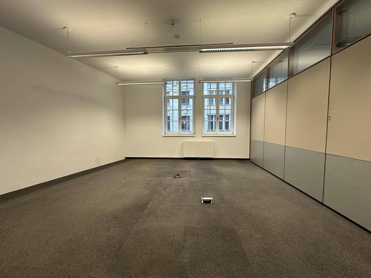 Leerer Büroraum mit Teppichboden, Fenstern und einer Wand mit integrierten Schränken, bereit zur individuellen Gestaltung.